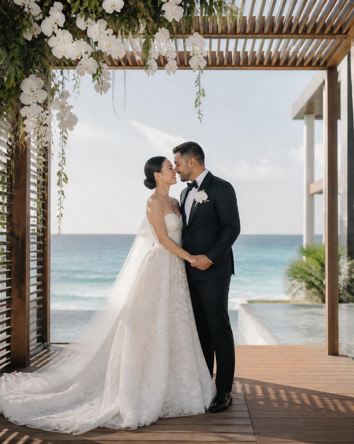An Asian bride and Latino groom under a modern wooden pergola with cascading orchids and sweeping Caribbean views in Riviera Maya